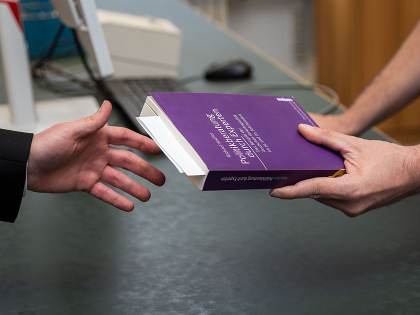 Buchübergabe Ein Bibliotheksmitarbeitender reicht einem Benutzer ein Buch.