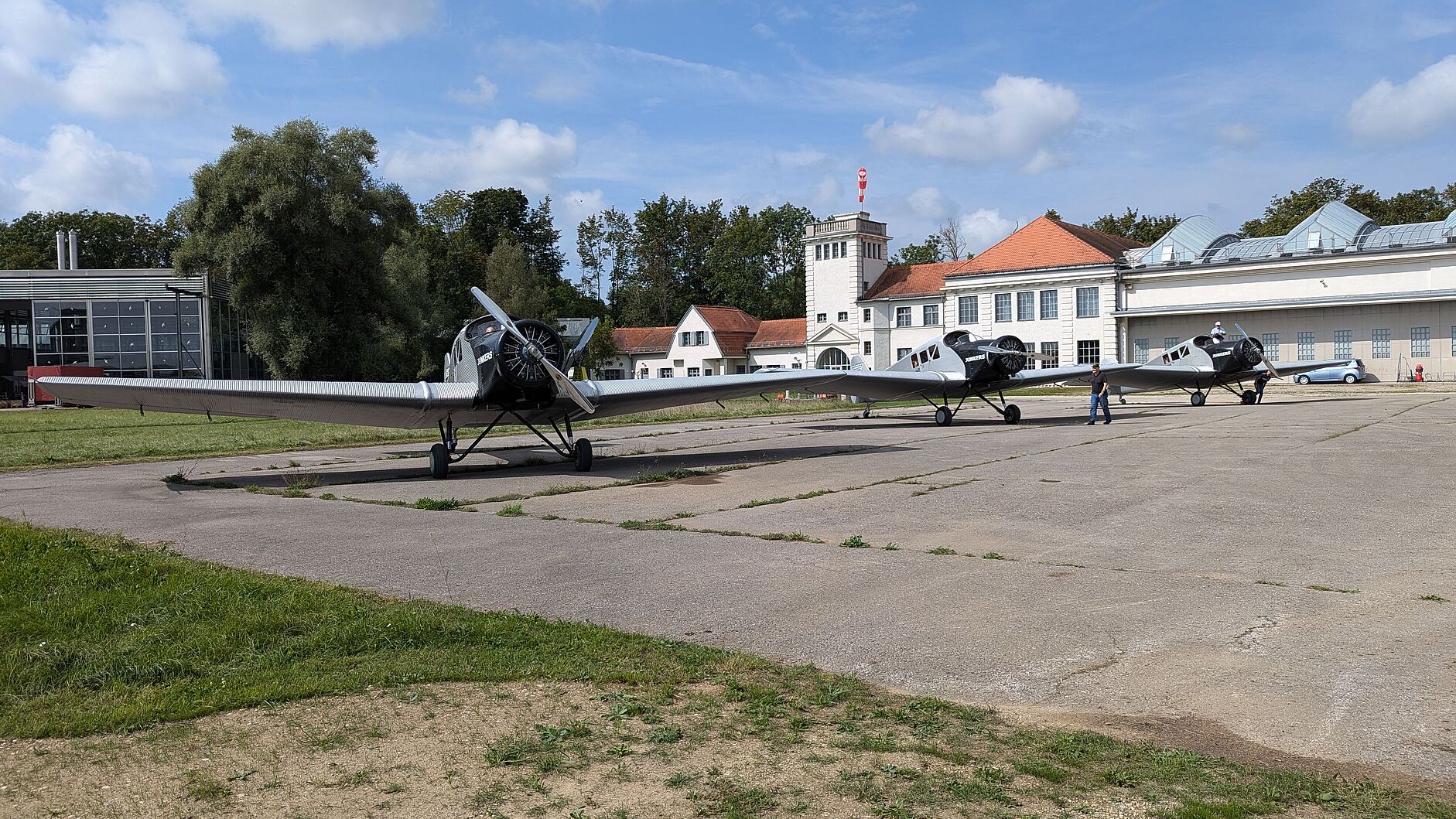 Drei silberne Propellermaschinen stehend auf Betonfläche. Längliches Gebäude mit Türmchen im Hintergrund.