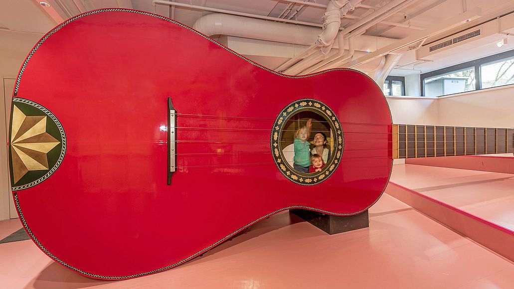 Riesengitarre Drei Kinder spielen mit der Riesengitarre. Zwei von ihnen sind in der Gitarre, ein Junge zupft von außen eine Seite.
