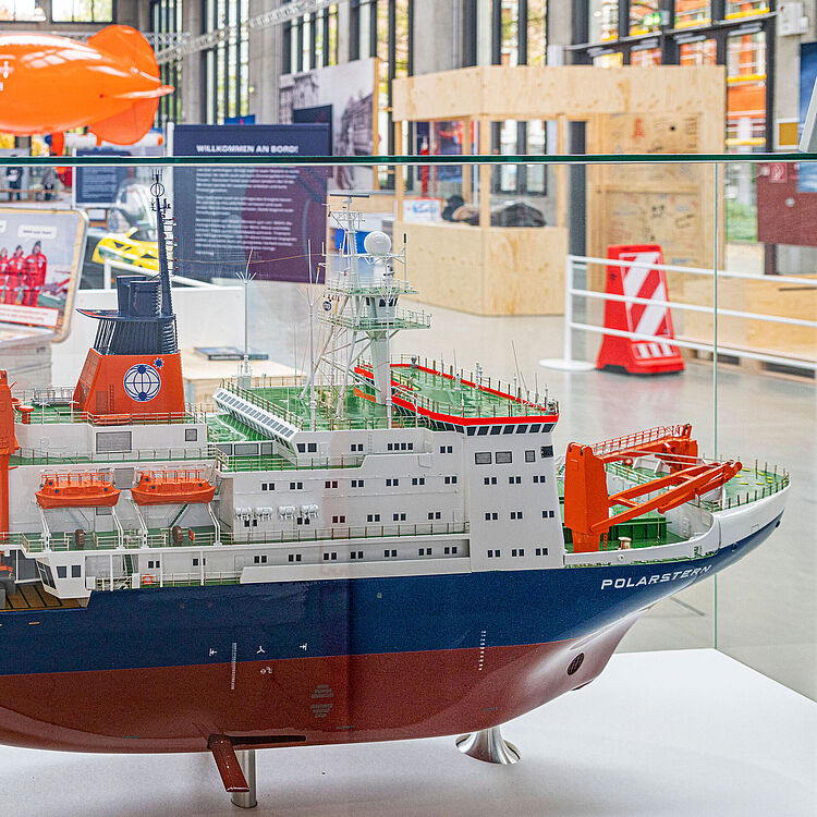 Ein Modell der POLARSTERN. Im Hintergrund sind der rote Heliumballon 'Miss Piggy', ein Schneemobil und weitere Exponate der Ausstellung sichtbar.