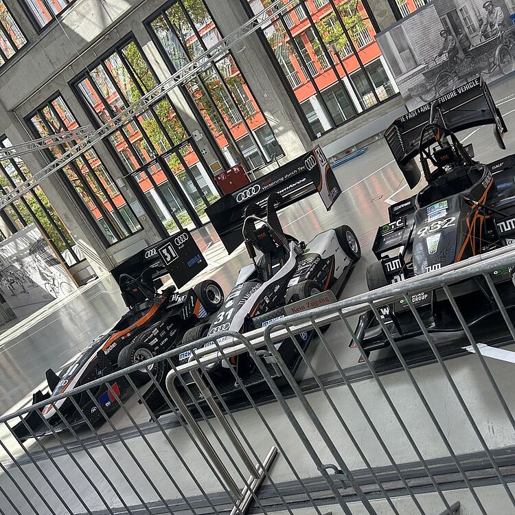 Drei offene Formelrennwagen im Ausstellungsraum einer Halle hinter Absperrgitter, schwarze und weiße Lackierung mit Sponsorenlogos (u. a. Audi, TUM), große Fensterfronten und rote Fassaden außen sichtbar.