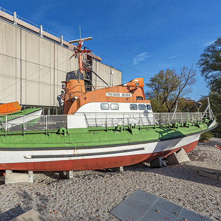 Rettungsboot „Theodor Heuss“ in Orange, Weiß und Grün auf Landgestell vor einem Betongebäude bei klarem Himmel, seitliche Ansicht mit Decksgeländer und Rettungsinseln.