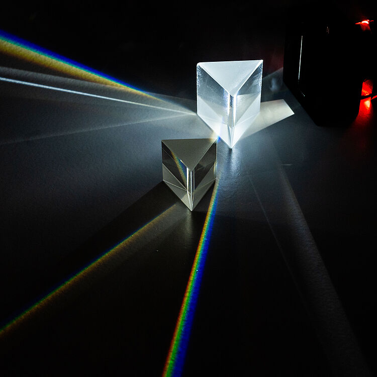 Zwei transparente optische Prismen auf dunklem Untergrund: ein größeres, leuchtend beleuchtetes Prisma rechts oben und ein kleineres vorne mittig, die Lichtstrahlen in scharfen weißen Linien und mehrfarbigen Regenbogenbändern brechen; im Hintergrund rechts ein schwarzes Gehäuse mit roten LED-Lichtern.