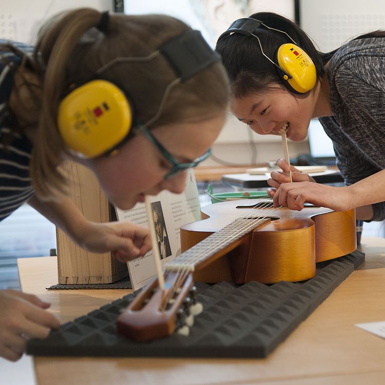Zwei junge Frauen mit Gehörschutz experimentieren mit einer Gitarre. Sie haben ein Holzstäbchen im Mund, welches an die Gitarre gehalten wird. Damit überprüfen sie die Schwingung der Gitarre, nach Zupfen einer Seite.