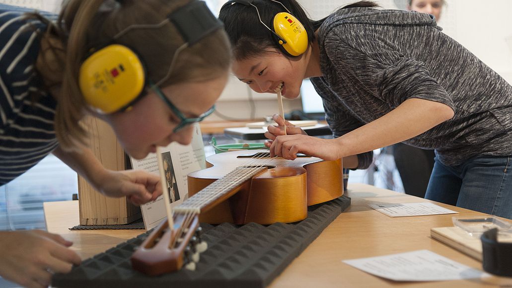 Hör-Werkstatt Zwei junge Frauen mit Gehörschutz experimentieren mit einer Gitarre. Sie haben ein Holzstäbchen im Mund, welches an die Gitarre gehalten wird. Damit überprüfen sie die Schwingung der Gitarre, nach Zupfen einer Seite.