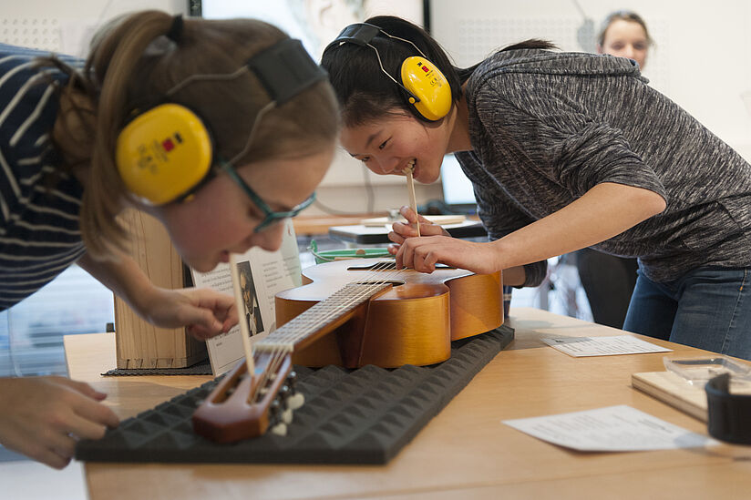 Hör-Werkstatt Zwei junge Frauen mit Gehörschutz experimentieren mit einer Gitarre. Sie haben ein Holzstäbchen im Mund, welches an die Gitarre gehalten wird. Damit überprüfen sie die Schwingung der Gitarre, nach Zupfen einer Seite.