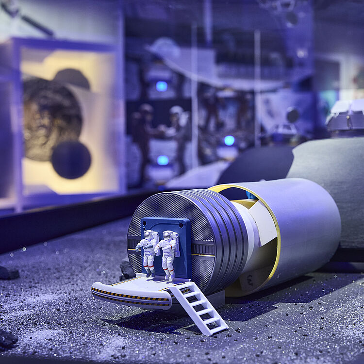 Miniaturmodell einer Mond- oder Planetenlandefähre auf grauem, staubigem Gelände mit zwei ausgestiegenen Astronautenfiguren auf einer kleinen Rampe, vor einem zylindrischen Modul und einem kuppelförmigen Habitat in einem musealen, blau beleuchteten Ausstellungsraum.