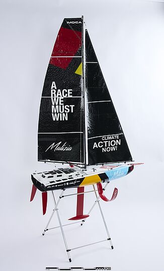 Modell eines Rennseglers auf Ständer mit schwarzem Großsegel, Schriftzug „A RACE WE MUST WIN“ links und „CLIMATE ACTION NOW!“ rechts, bunte Rumpfbemalung und roter Kiel.