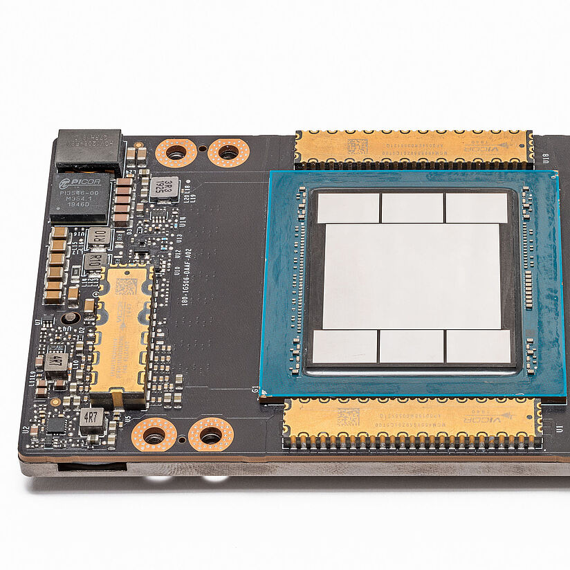 Fotografie eines Chips in Draufsicht. In der Mitte der Platine befinden sich silberne Flächen, von blauen Elementen umrandet. Am linken und rechten Rand befinden sich verschiedene Schaltelemente verschiedener Größe in Gold- und Grautönen.