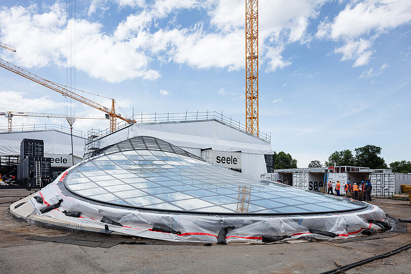 Blick von der Oberfläche auf das Dach der Bahnsteighalle. Ein großes, nahezu kreisrundes, fast flaches Fensterelement wurde auf die Öffnung einer Kelchstütze gebaut. Außen herum ist Baustelle. Auch am Lichtauge selbst sind noch Abdeckfolien angebracht.