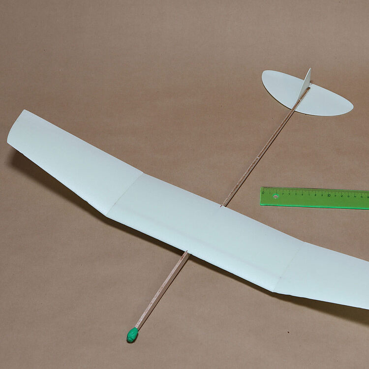 Ein handgebautes, weißes Styropor-Pfeilflugzeug (Gleiter) mit langer dünner Holmstange, ovalem Höhenleitwerk hinten und grünem Modellier-Knetboden am Bug auf braunem Papieruntergrund neben einem grünen 30-cm-Lineal.