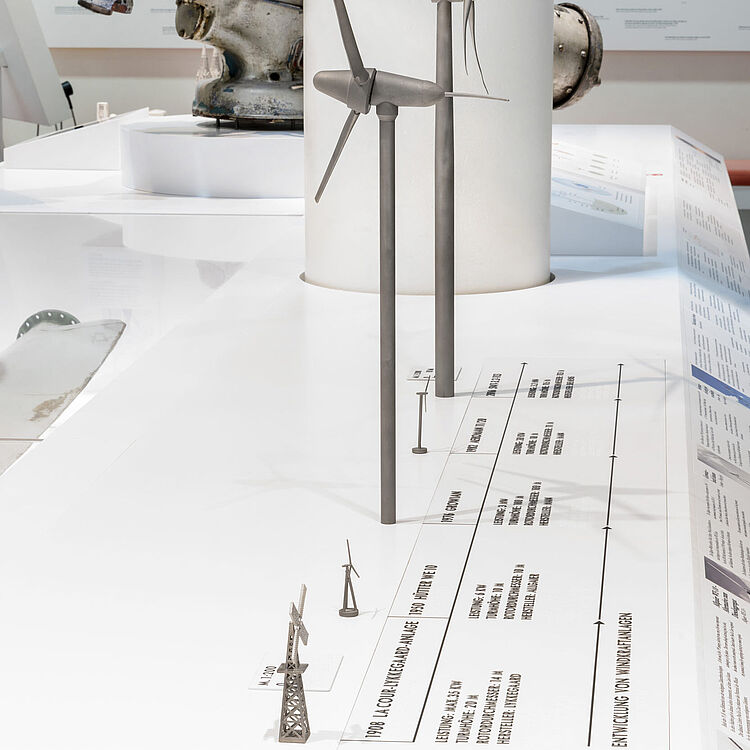 Tast-Modelle mehrerer Windräder: vier graue Turmmodelle mit Rotorblättern unterschiedlicher Bauarten auf einer weißen Präsentationsfläche.