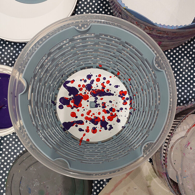 Draufsicht auf eine Salatschleuder auf einer gepunkteten Basteltischdecke, gefüllt mit roten und violetten Farbklecksen auf einer Papiereinlage.