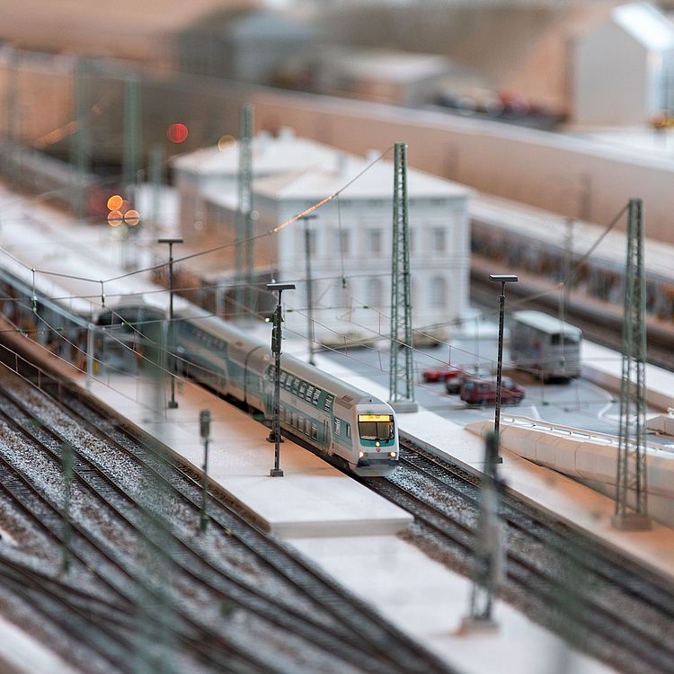 Detail der Modelleisenbahn. Zu sehen sind der Bahnhof mit Parkplatz, zwei überdachte Bahnsteige und ein einfahrender Zug.
