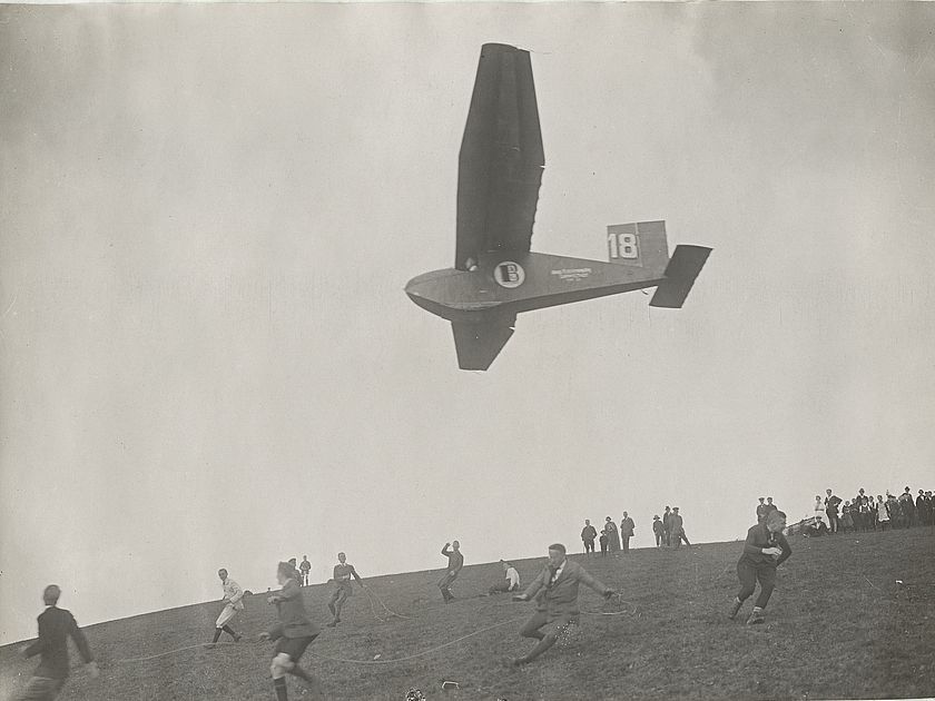 Ein Segelflugzeug hat sich an einem Hang in die Luft erhoben, unten sind mehrere Männer mit den Seilen beschäftigt.