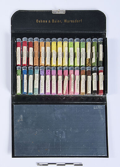 Geöffnetes schwarzes Etui mit der Goldprägung "Oehme & Baier, Warnsdorf" im Deckel, innen zwei Reihen kleiner Glasröhrchen mit farbiger Granulatfüllung in Regenbogenfarben (Rot, Orange, Gelb, Grün, Blau, Violett, Rosa, Braun, Schwarz) jeweils mit weißen Beschriftungszetteln, darunter eine blanke Spiegel- oder Metalltafel.