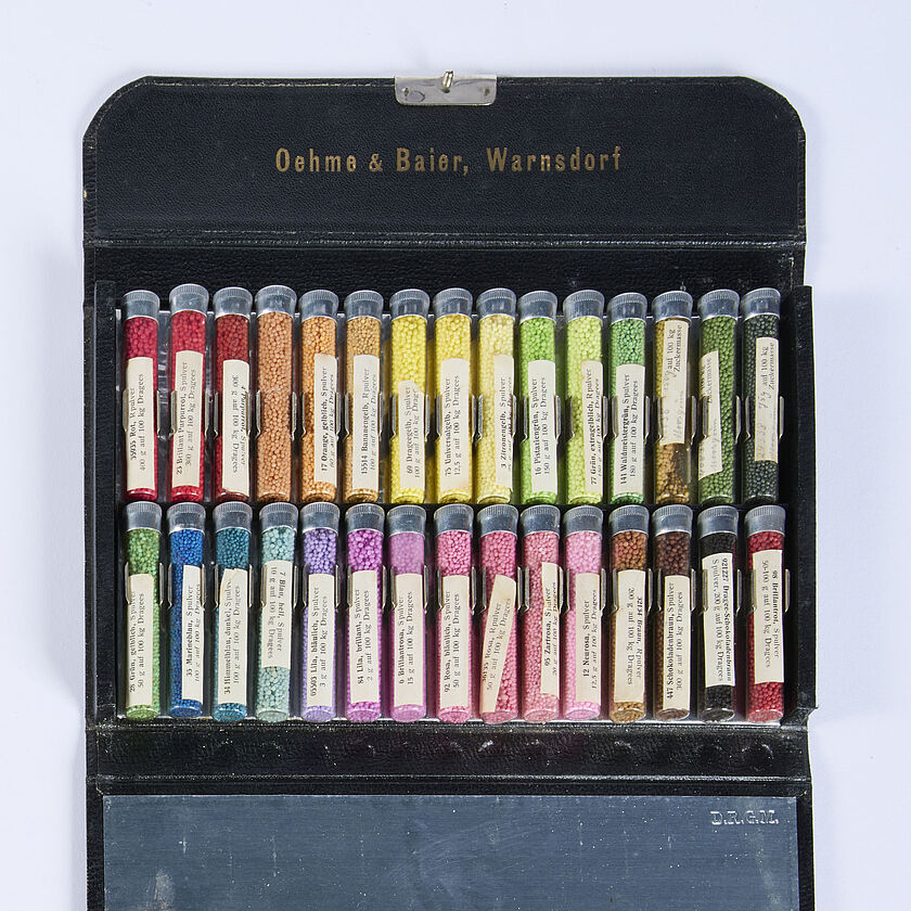 Geöffnetes schwarzes Etui mit der Goldprägung "Oehme & Baier, Warnsdorf" im Deckel, innen zwei Reihen kleiner Glasröhrchen mit farbiger Granulatfüllung in Regenbogenfarben (Rot, Orange, Gelb, Grün, Blau, Violett, Rosa, Braun, Schwarz) jeweils mit weißen Beschriftungszetteln, darunter eine blanke Spiegel- oder Metalltafel.