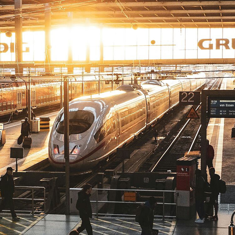 Die Bahnsteighalle des Münchner Hauptbahnhofs bei Abendstimmung. Ein ICE-Zug wartet auf seine Abfahrt. Auf den Bahnsteigen sind Reisende unterwegs.