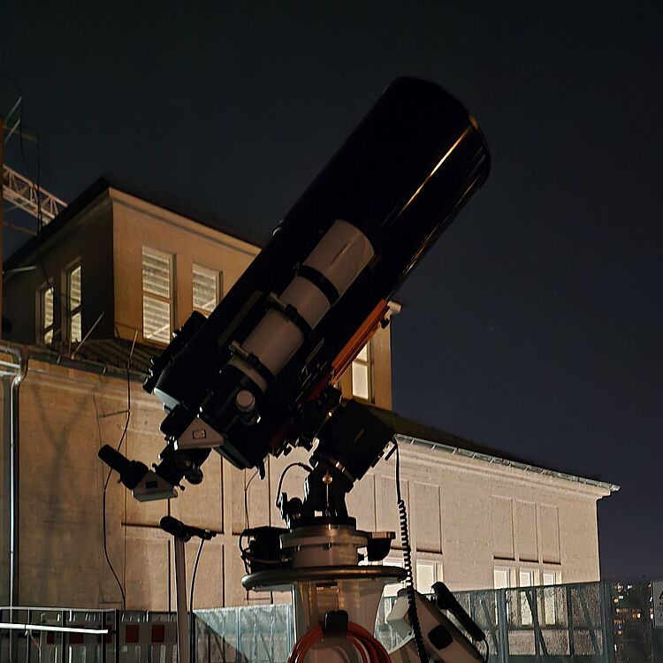 Ein großes, montiertes Spiegelteleskop auf einem weißen mobilen Rollwagen auf der Dachterrasse des Deutschen Museums bei Nacht.