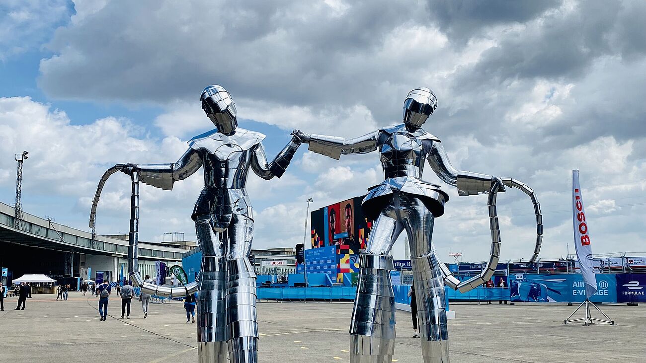 zwei Menschen auf Stelzen verkleidet als Aliens in silbernen Outfits.