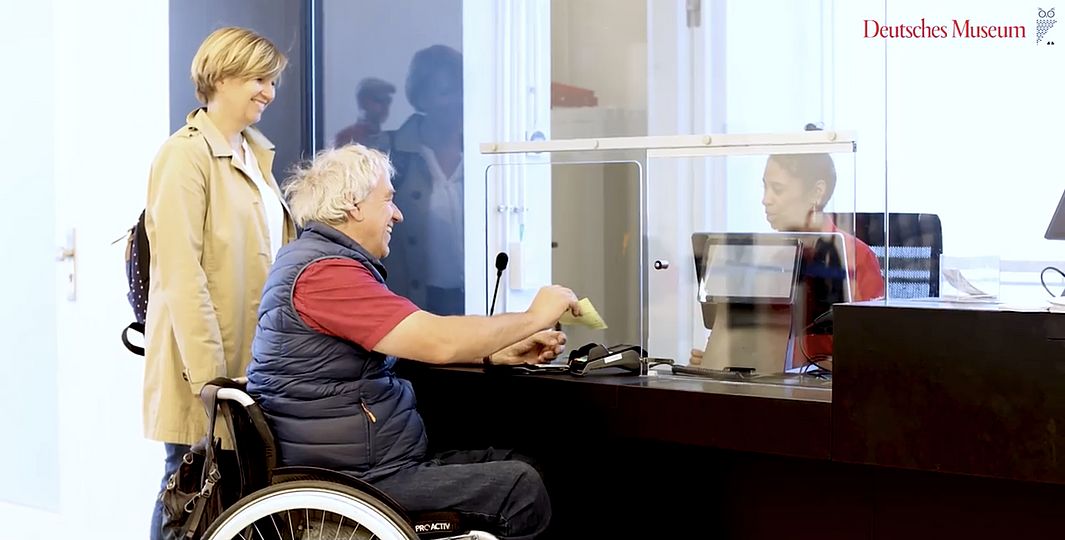 Besucher im Rollstuhl und Begleiterin kaufen Tickets am Ticketschalter des Deutschen Museum.
