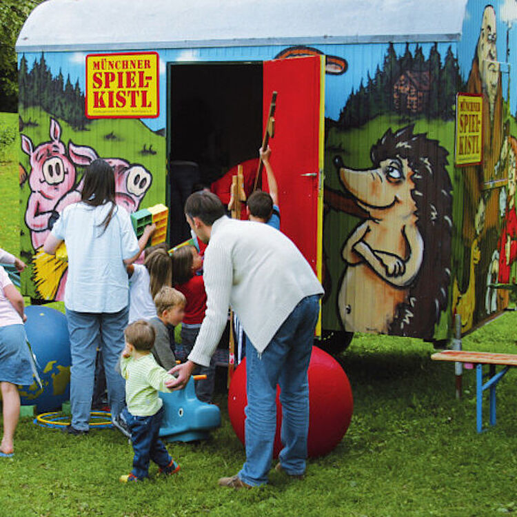 Eine bunte Spielwagen‑Szene auf einer Wiese: ein bemalter Anhänger mit geöffneten roten Türen und der Aufschrift "Münchner Spielkistl", vor dem Kinder und Erwachsene bunte Spielgeräte ausladen — große Gymnastikbälle, Bauklötze, Holzspielzeug und ein blaues Rutschpferd; im Hintergrund Bäume und grünes Gras.