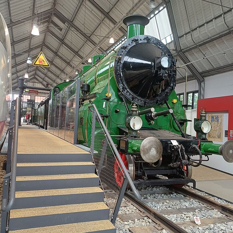 Grün lackierte Dampflokomotive S 3/4, frontal rechts sichtbar mit großem schwarzem Schornstein, runden Scheinwerfern, roten Speichenrädern und Gleisen; links führt eine Treppe mit Geländer zu einer erhöhten Besucherplattform, im Hintergrund Hallendach mit Leuchten und ein gelbes Warnschild.