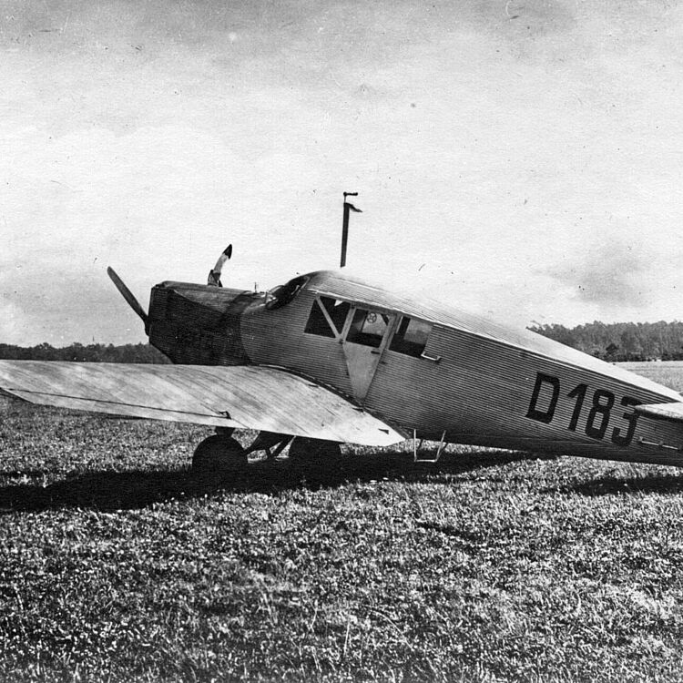 Schwarzweiß-Foto eines einmotorigen Landflugzeugs mit Kennung "D183" auf dem Rumpf, geparkt auf einer Grasfläche; Seitenansicht zeigt einen starren Tiefdecker mit zweiblättriger Holzpropellernase, geschlossener Kabine mit rechteckigen Fenstern, spitz zulaufendem Heckleitwerk und sichtbaren Reifen im Fahrwerk vor einer baumbestandenen Horizontlinie.