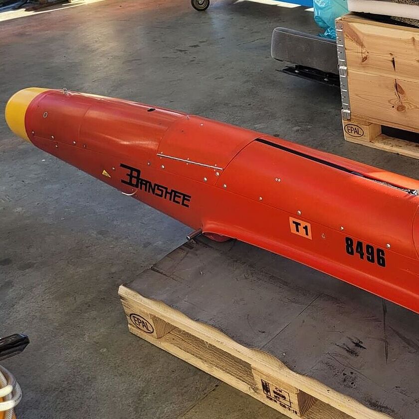 Totale einer Zieldarstellungsdrohne in einem Lagerraum, im Hintergrund Holzkisten auf Paletten. Die Drohne im Vordergrund ist zylinderförmig-länglich, hat eine gelbe abgerundete Spitze und ein Einschlussloch am Leitwerk. Sie wird auf einer Palette gelagert. Die Aufschrift "Banshee" auf dem vorderen Teil und "Qinteiq" auf dem Leitwerk sind gut lesbar.