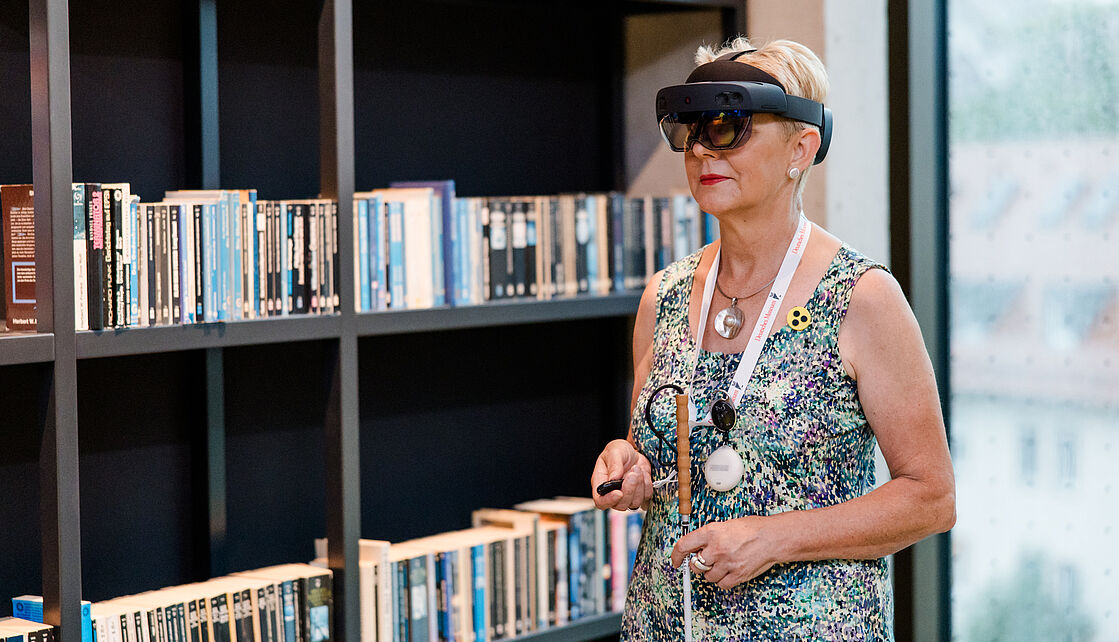 HoloAudioTour durchs Zukunftsmuseum Durchs Zukunftsmuseum mit der HoloAudioTour. Das Bild zeigt eine Person mit einer HoloLens Datenbrille in der Bibliothek des Museums. Die Person hält in einer Hand einen Blindenstock und in der anderen Hand einen Controller für die Datenbrille.