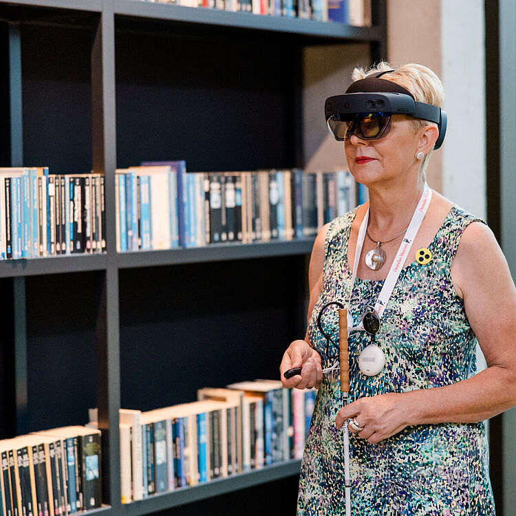 Durchs Zukunftsmuseum mit der HoloAudioTour. Das Bild zeigt eine Person mit einer HoloLens Datenbrille in der Bibliothek des Museums. Die Person hält in einer Hand einen Blindenstock und in der anderen Hand einen Controller für die Datenbrille. 