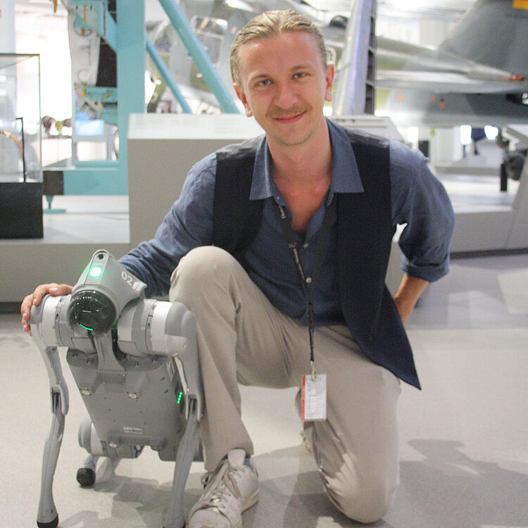 Museums-Communicator Florian kniet neben seinem Roboter-Hund. Im Hintergrund sind Flugzeuge aus der Modernen Luftfahrt-Ausstellung zu erkennen.