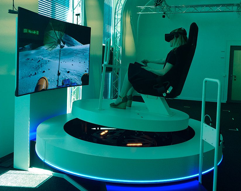 Foto des Fahrsimulators im VRlab mit einer Person auf dem Simulator mit VR-Brille. Auf dem Bildschirm sieht man die Mondoberfläche.