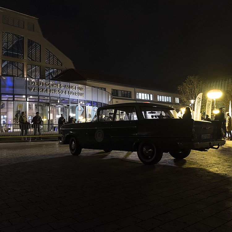 Nachtszene vor dem Gebäudeeingang des Deutschen Museums Verkehrszentrum: im Vordergrund die Silhouette eines schwarzen Oldtimer-Kombis auf gepflastertem Platz, im Hintergrund beleuchtete Glasfassade mit Menschen, Banner und weitere klassische Fahrzeuge unter warmen Straßenlaternen.
