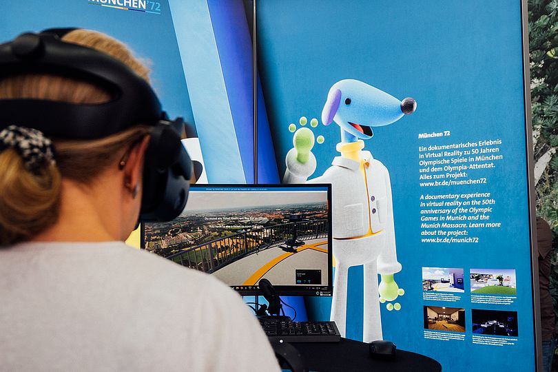 Foto einer Person mit einer VR-Brille von hinten. Der Blick über die Schulter zeigt einen Bildschirm mit Inhalten der München 72 Anwendung sowie weitere Informationen dahinter.