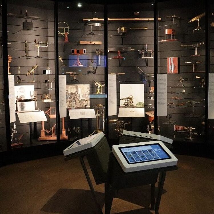 Museumsvitrine mit zahlreichen historischen Messinstrumenten und optischen Geräten an den Wänden, davor zwei interaktive Touchscreen-Pulte auf Ständern, gedimmte Beleuchtung.