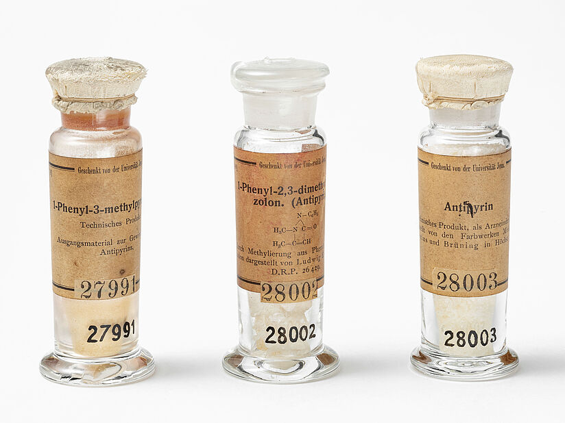 Drei schmale, gläserne Apothekerflaschen mit vergilbten Labels und aufgebrachten Inventarnummern.