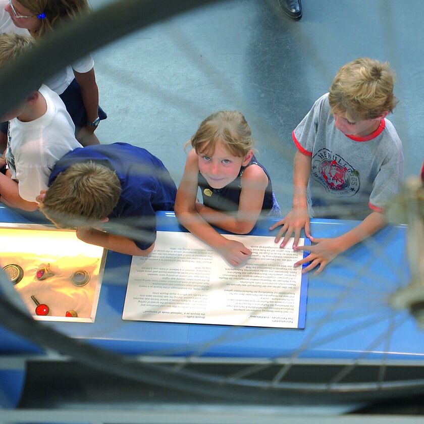Vogelperspektive auf eine Gruppe von Kindern, die an einem blauen Ausstellungstisch lesen und Experimente betrachten; im Vordergrund unscharf ein Fahrrad-Rad mit Speichen, am Tisch Infotafeln und kleine Objekte in einer beleuchteten Vertiefung.