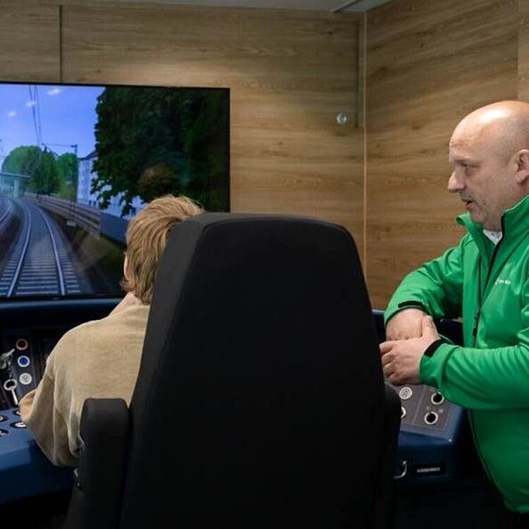 Personen sitzen in einem mobilen S-Bahn-Simulator, ein Ausbilder erklärt das virtuelle Zugfahren auf einem großen Bildschirm.