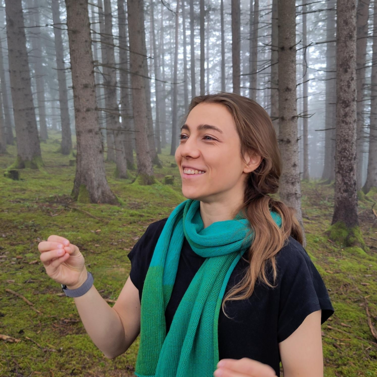 Eine lächelnde Frau mit braunem Haar und grün-türkisem Schal über einem schwarzen T-Shirt steht in einem nebligen Nadelwald mit moosigem Boden und hohen, schlanken Baumstämmen, die Hände leicht erhoben, Blick nach rechts.