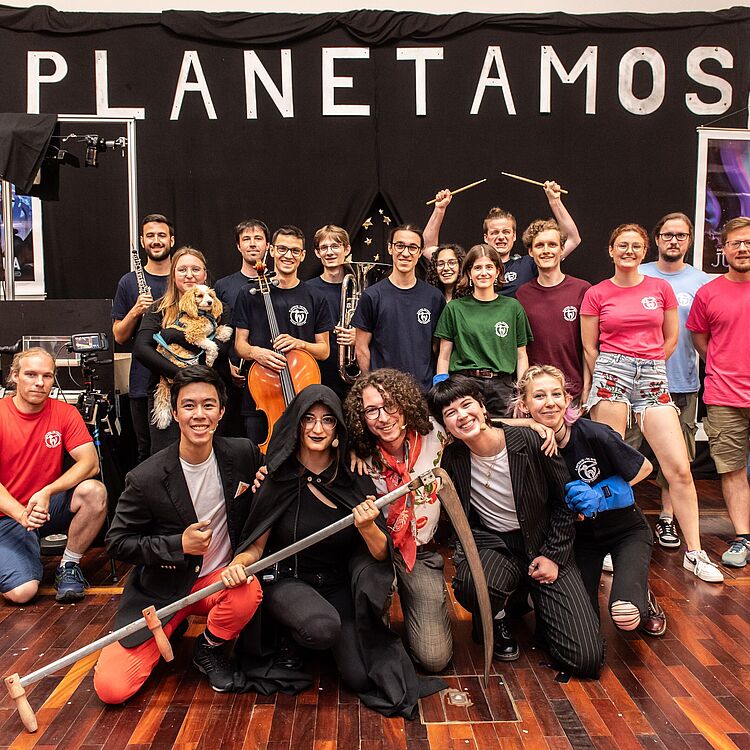 Die Studierenden des Physikshow-Musicals haben sich für ein Gruppenfoto aufgestellt. Neben einigen Kostümen und Requisiten sind auch Musikinstrumente zu sehen. Die Studierenden der Physikshow-Musical-Gruppe