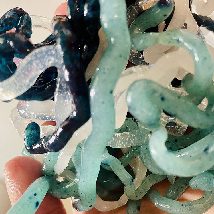 Nahaufnahme von mehreren miteinander verschlungenen, gläsern wirkenden Bioplastik-Schnüre aus Algenin Blau-, Türkis- und Opalweißtönen mit glänzender, leicht transparent strukturierter Oberfläche, die in einer Hand gehalten werden.