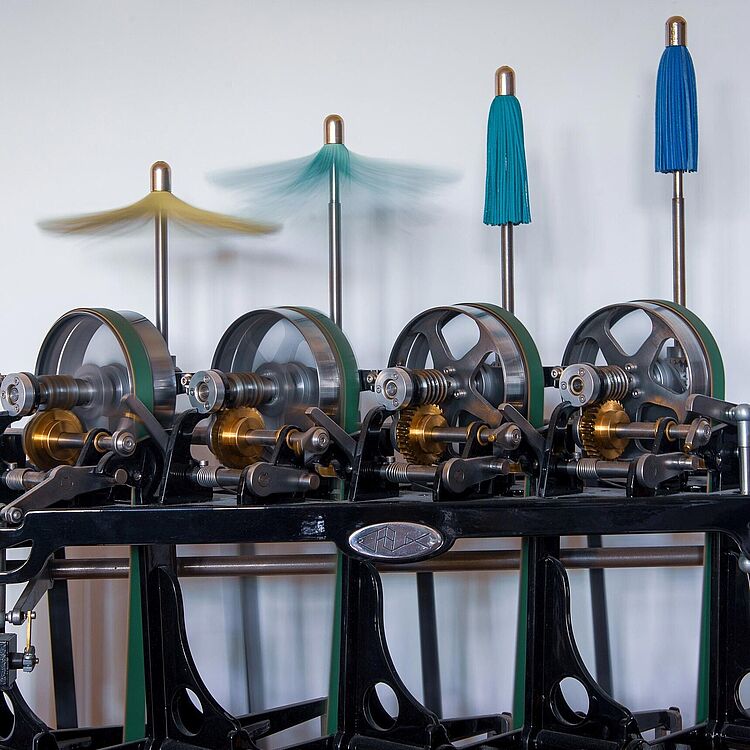 Eine historische industrielle Maschine mit vier parallel angeordneten Schwungrädern, sichtbaren Zahnrädern und Metallachsen auf einem schwarzen Gestell; oben sind vier rotierende bürstenartige Pinselaufsätze in Gelb, Hellblau und zwei Blau­tönen.