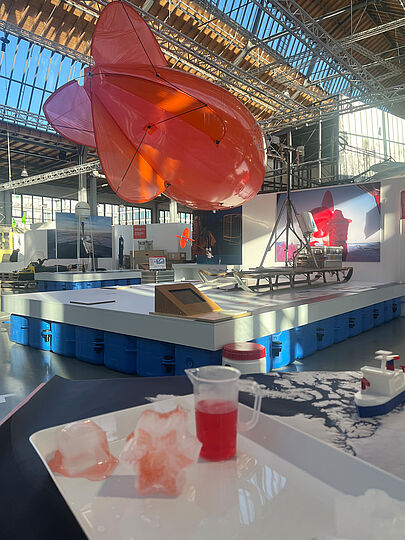Sonderausstellungsfläche mit einer großen, roten Heliumballon „Miss Piggy“. Im Vordergrund stehen eine weiße Platte mit schmelzendem Eis und eine rote Flüssigkeit in einem transparenten Messbecher. Im Hintergrund sind weitere Exponate und ein Modell eines Schlittens zu sehen.