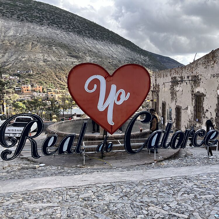 Großes rotes Herz-Skulptur mit weißer Schrift "Yo" über dekorativem Schriftzug "Real de Catorce" auf gepflastertem Platz vor verfallener Lehm-Mauer, Berg im Hintergrund und zwei Personen.