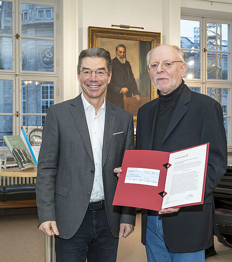 Zwei Männer mit einer Urkunde in den Händen vor dem gemalten Porträt von Oskar von Miller