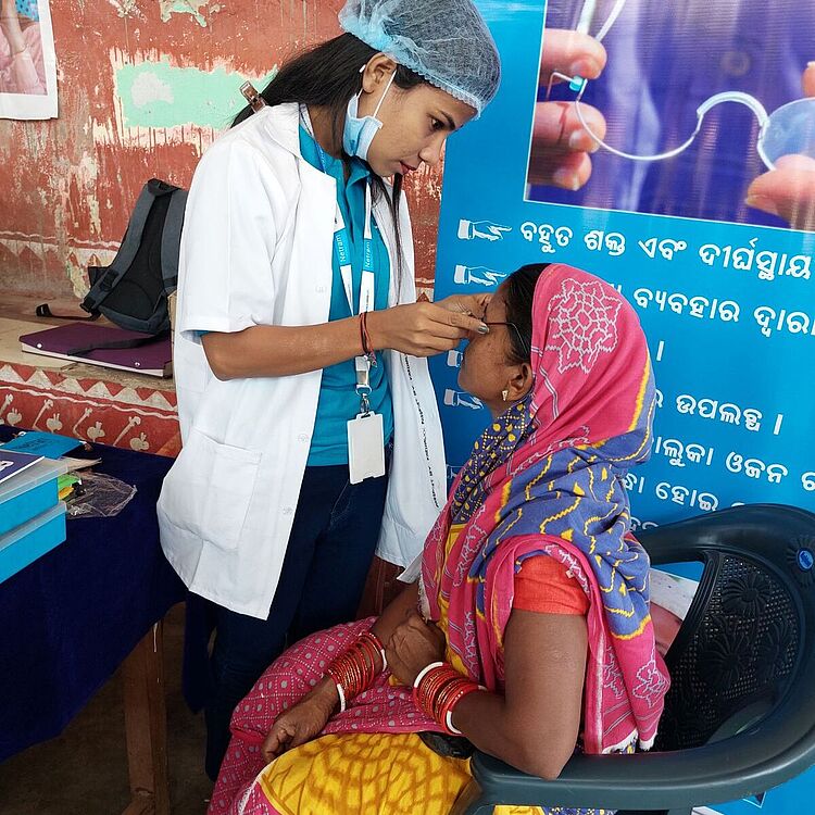 Eine Gesundheitshelferin in weißem Kittel und Haarnetz setzt einer sitzende Frau in buntem Sari eine Brille auf.