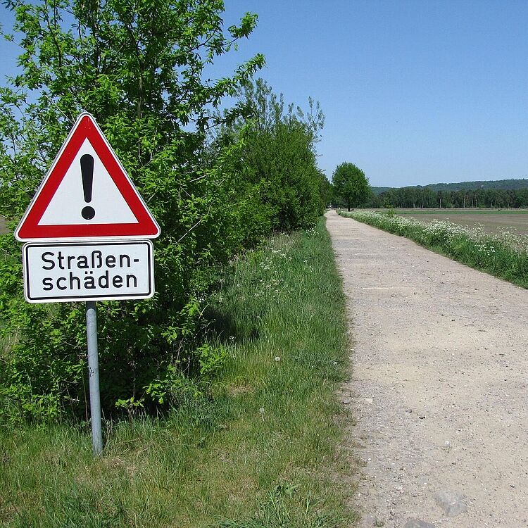 Ländlicher Schotterweg unter blauem Himmel, links ein Warnschild mit rotem Dreieck und Ausrufezeichen und Zusatztafel „Straßenschäden“, Bäume und Feldgrenzen entlang des Wegs.