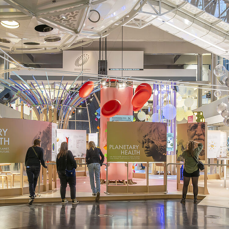 Ausstellungshalle mit dem thematischen Messestand "PLANETARY HEALTH": bunte Installationen, rote Hutschalen hängend, Informationswände, leuchtende Kugeln und vier Besucherinnen die Exponate betrachten.