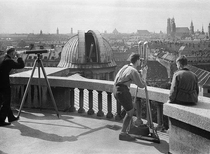 Besucher blicken auf der großen Terrasse des Deutschen Museums durch zwei Fernrohre in Richtung der Münchner Innenstadt. Im Hintergrund ist die geöffnete Westkuppel mit dem Zeiss-Refraktor zu sehen. Das Foto ist vor 1945 entstanden.